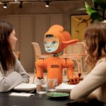 Two women talk to an orange robot at a table.
